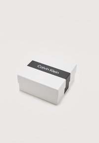 Λευκό ορθογώνιο κουτί με μαύρη λωρίδα όπου είναι τοποθετημένο το λογότυπο "Calvin Klein" στο κέντρο της λωρίδας, σε φόντο ανοιχτού γκρι.