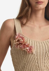 Beige stickad linne med breda axelband, prydd med en klunga rosa blomdetaljer på vänster axel. Texturerad tyg med ribbat mönster.