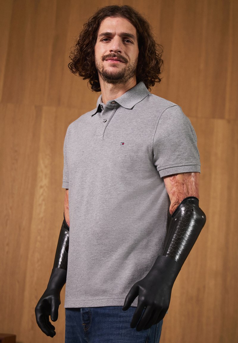 Grå poloshirt med en liten logo, blå jeans, og svarte proteseforrearms med teksturerte og glatte overflater. Trebakgrunn.