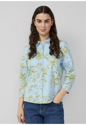 Kobieta o długich, ciemnych kręconych włosach, ubrana w jasnoniebieską kwiecistą koszulę na guziki i niebieskie dżinsy, stojąca z jedną ręką w kieszeni.