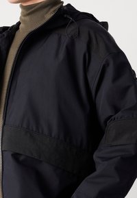 Tmavě modrá bunda s hladkým povrchem, černými detaily, kapucí a zapínáním na zip. Texturované pásy na ramenou dodávají designu zajímavý detail.