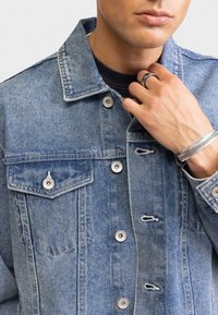 Giacca in denim azzurro chiaro con due tasche sul petto, bottoni in argento e un leggero sbiadimento. Presenta un colletto classico e leggeri sfilacciamenti sui bordi.
