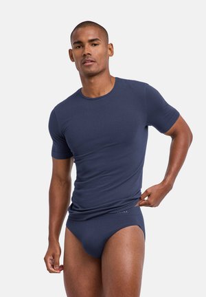 Mann trägt eng anliegendes dunkelblaues T-Shirt und passende Slips, richtet den Bund mit einer Hand vor einfarbigem Hintergrund.