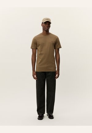 T-shirt en coton marron à manches courtes, portant un petit logo sur la poitrine gauche ; associé à un pantalon en denim foncé à jambe droite et des chaussures noires.