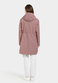 Ljusrosa, lättviktig kappa med huva och elastisk snörning i midjan. Har långa ärmar och rak skärning, bärs över vita byxor.