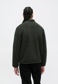 Dark green fleece jacket with a high collar and relaxed fit, featuring a textured surface and ribbed hem, paired with black pants.