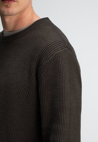 Dunkelbrauner, gerippter Strickpullover mit Rundhalsausschnitt und strukturiertem Stoff. Zeigt Details an der Schulternaht. Keine sichtbaren Marken oder Muster.