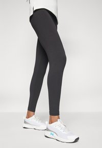 Fekete sportleggings fehér és szürke sportcipőkkel, amelyek hálós részletekkel és világoskék akcentusokkal rendelkeznek. Simább, rugalmas szövettextúra.