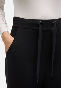 Pantalones negros con cintura ajustable, que cuentan con dos bolsillos laterales y una textura de tela suave. Los cordones tienen detalles metálicos negros.