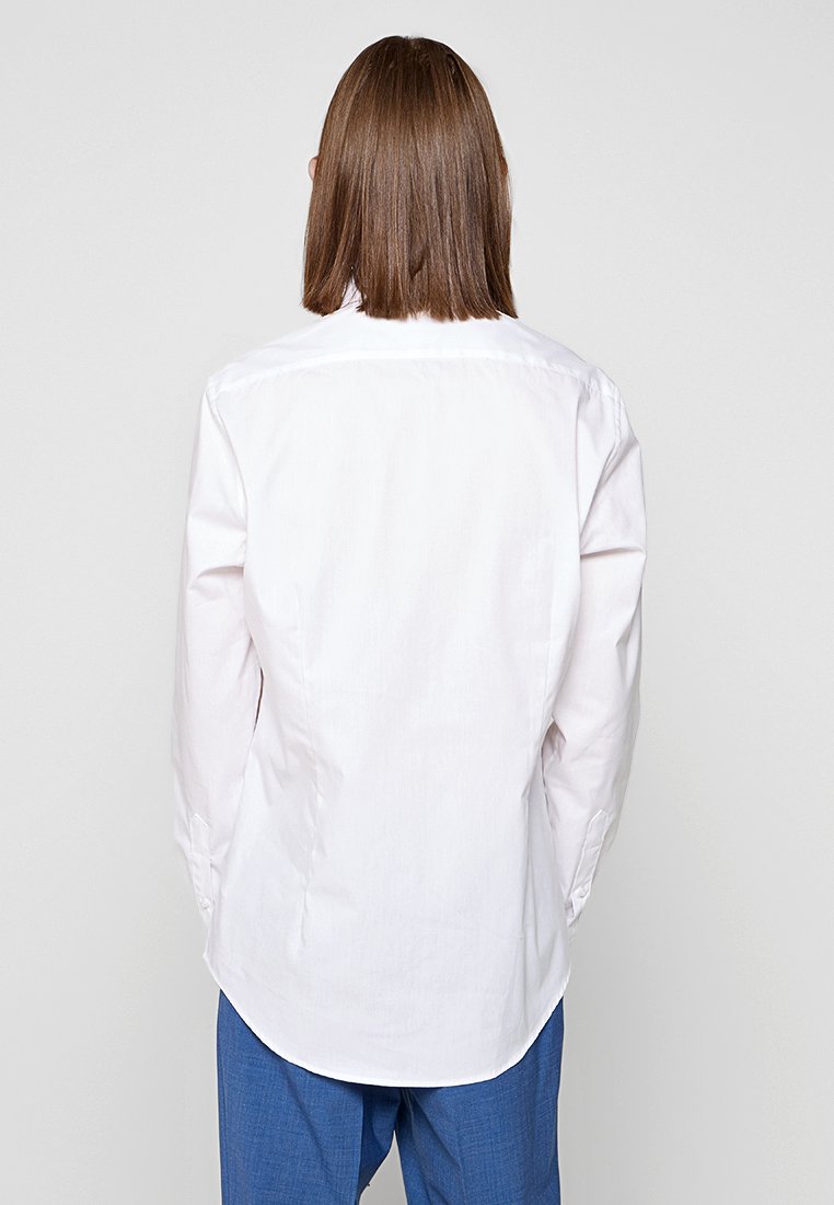 Weißes Hemd mit geradem Schnitt, langen Ärmeln, abgerundetem Saum und Rückenpasse. Getragen mit einfarbigen blauen Hosen.