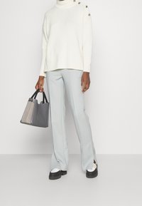 Vit stickad polotröja med knappdetaljer på ena axeln, i kombination med ljusblå skräddarsydda byxor och en flerfärgad handväska.