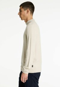 Joven en suéter de punto beige de perfil contra un fondo blanco, vistiendo pantalones oscuros y con una postura relajada.