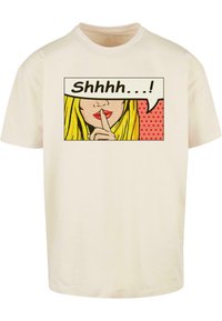 Beige bomull t-skjorte med et trykt pop art-design som viser ansiktet til en kvinne, som gir en stillhetsgest, svart tekst "Shhh...." og en rød prikkete bakgrunn.