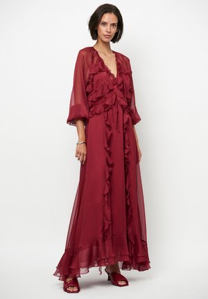Longue robe bordeaux en tissu transparent avec des accents de volants, un décolleté plongeant en V et des manches longues. Assortie à des chaussures à brides assorties.