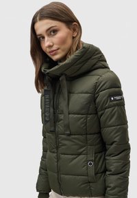 Olijfgroene pufferjack met een hoge kraag, horizontale stiksels, een rits aan de voorkant en een logo-opdruk op de mouw. Bevat een ritszak.