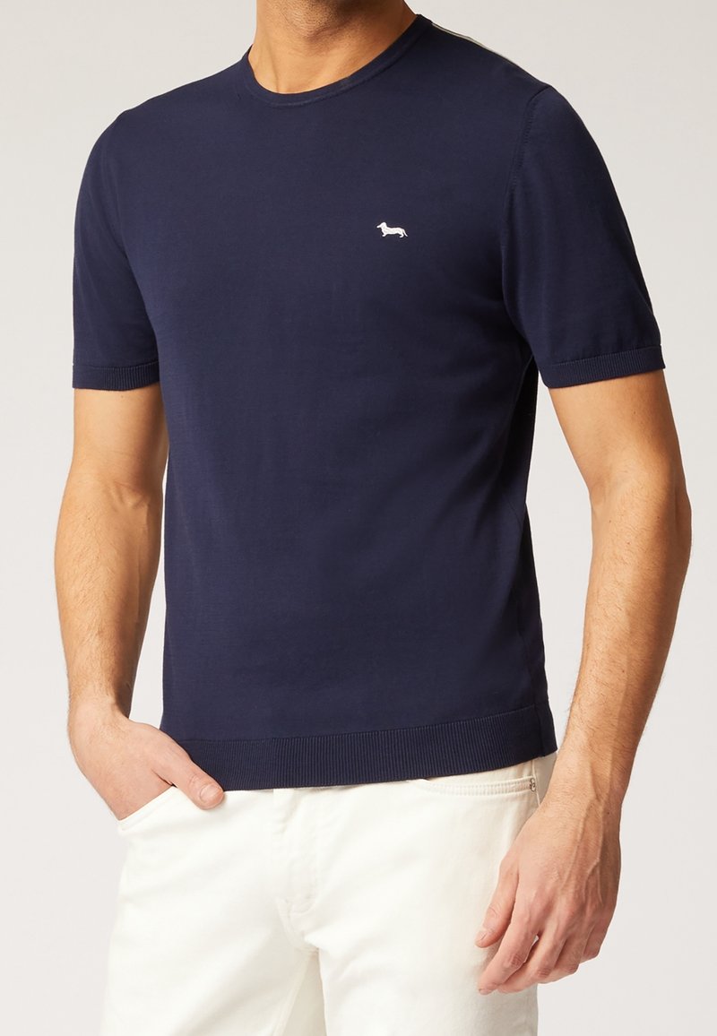 Mann trägt ein marineblaues, kurzärmliges Strickhemd mit einem kleinen weißen Dackel-Logo auf der Brust und weiße Hosen.