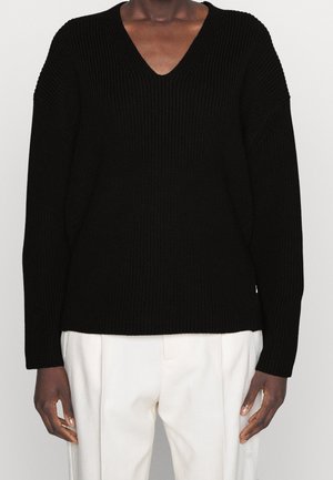 Homme portant un pull en maille noir à col en V avec manches longues, associé à un pantalon blanc plissé, debout devant un fond uni.