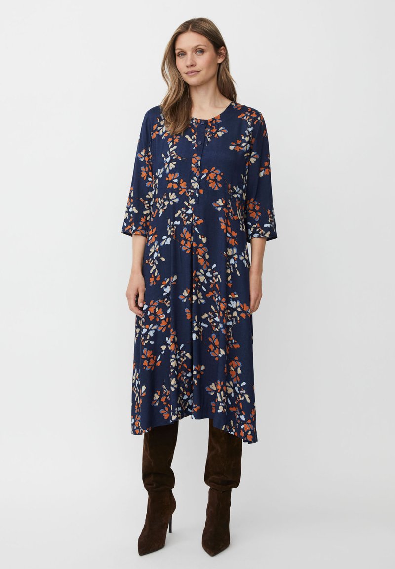 Mörkblå klänning med blommönster i orange, vitt och grönt. Har trekvartsärmar, rund halsringning och ett luftigt, flödande snitt.