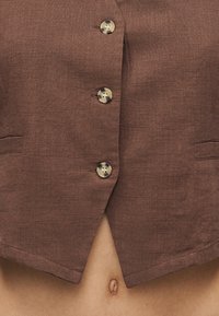 Καφέ υφασμάτινο σακάκι με τρεις κουμπιά από χελωνόκρανο, ελαφρώς ανοιχτό στο κάτω μέρος, αποκαλύπτοντας έναν γυμνό μέση με ορατή τον αφαλό.