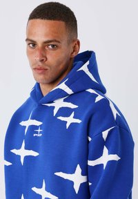 Sudadera azul con grandes patrones de estrellas blancas, un bolsillo canguro frontal y capucha con cordón. Hecha de tela suave y gruesa.