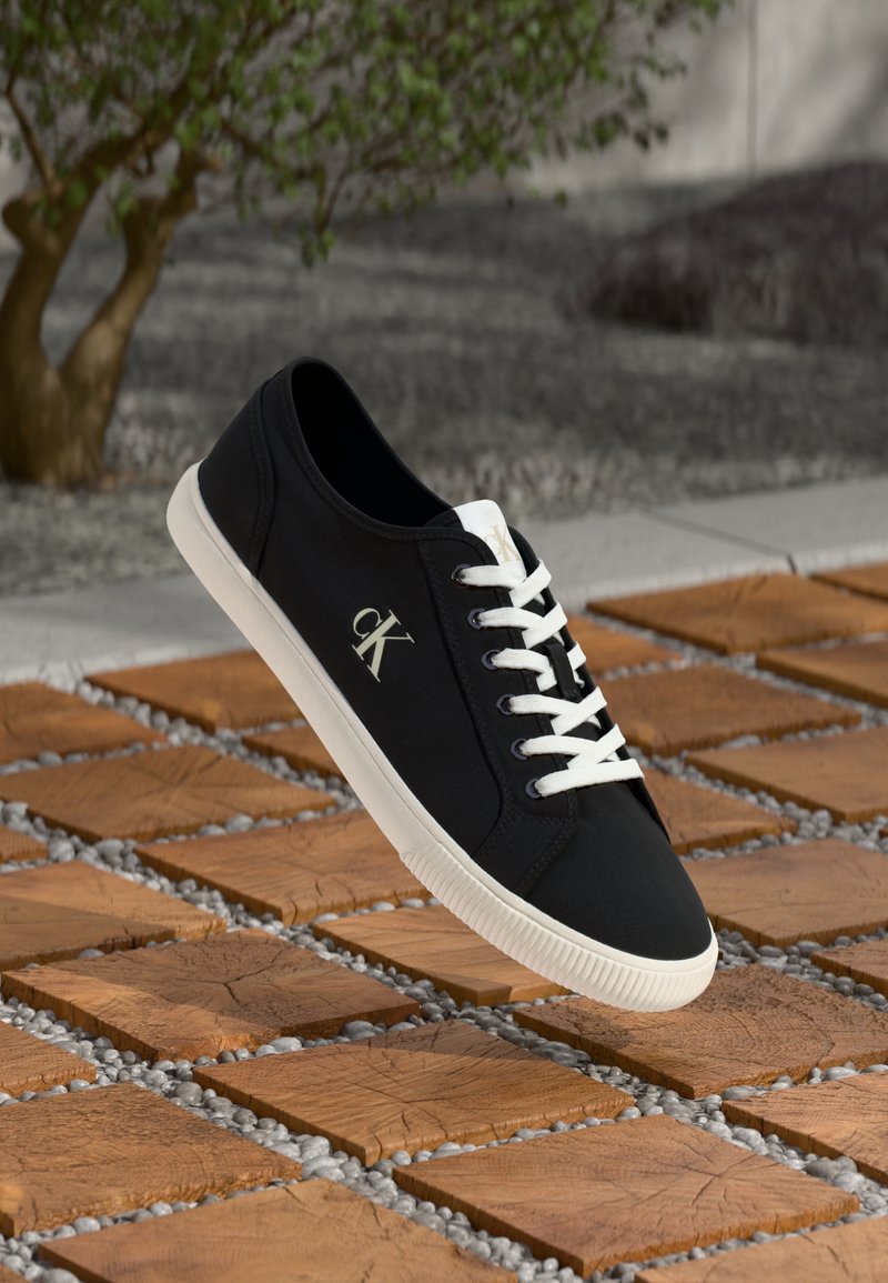 Fekete vászon sneaker fehér fűzőkkel és oldalán a "cK" logóval, fából készült négyzet alakú csempék felett, kisméretű kaviccsal az ívek között.