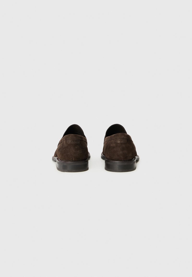 Mocassins en daim marron avec un petit talon, des orteils ronds et des détails de couture minimaux. La vue arrière met en avant la texture douce et les lignes de conception épurées.