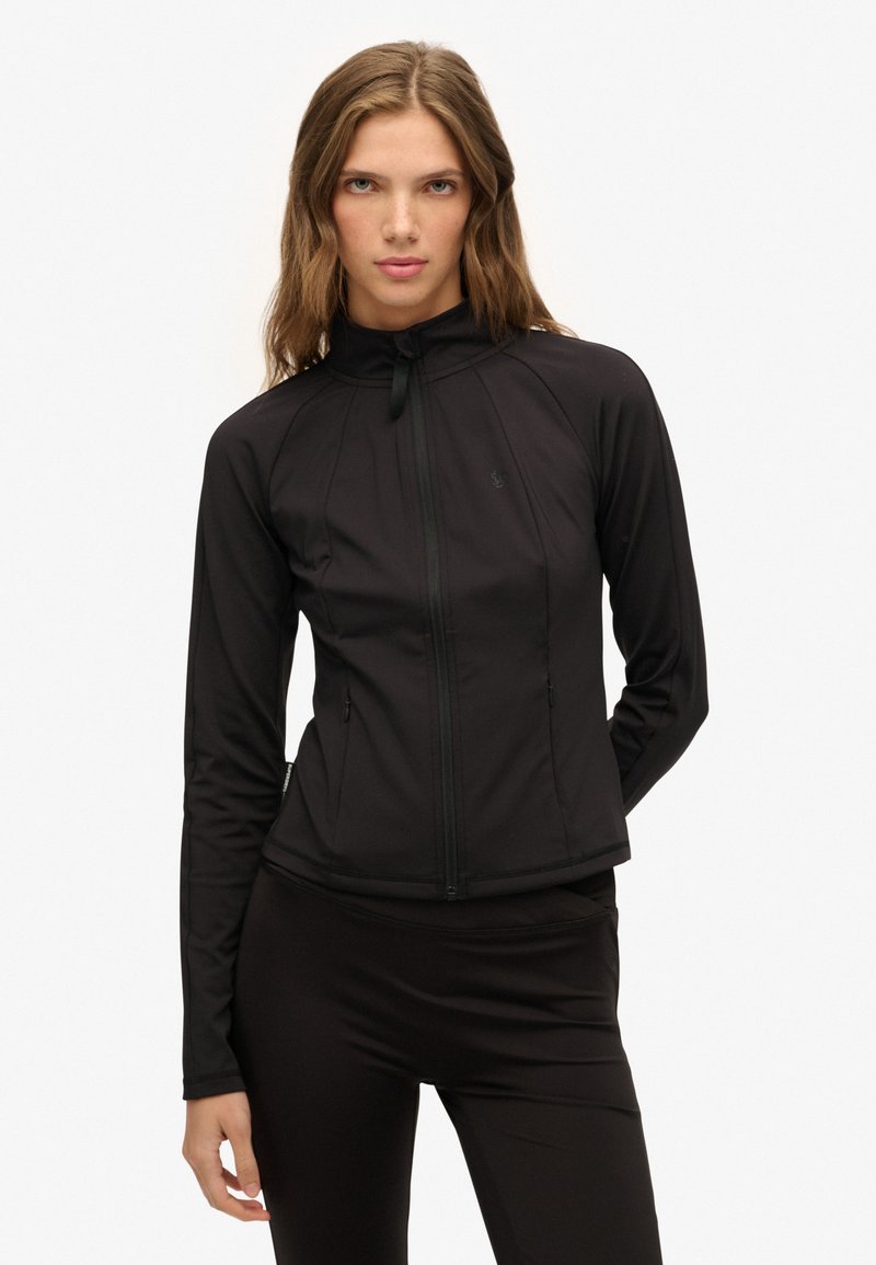 Veste de sport noire à col montant, fermeture éclair intégrale et deux poches latérales. Fabriquée en matériau lisse et flexible, avec des accents ton sur ton discrets.