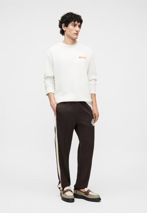 Νέος άνδρας με κατσαρά μαλλιά που φορεί λευκό πουκάμισο με μακριά μανίκια, σκούρο καφέ παντελόνι με πλευρική ρίγα και μπεζ μοκασίνια με λεπτομέρεια αλυσίδας.