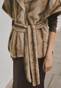 Faux fur vest in soft brown tones, featuring a wide collar, belt tie, and textured pattern. Worn over a long-sleeve brown garment.