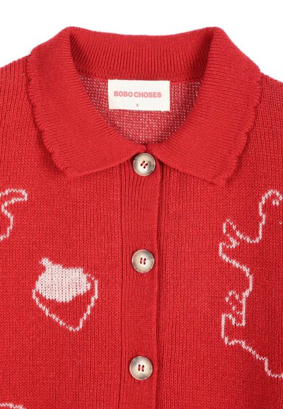 Cardigan rouge en tricot avec un col arrondi, trois boutons beige et des formes abstraites blanches sur le tissu. L’étiquette indique BOBO CHOSES taille S.