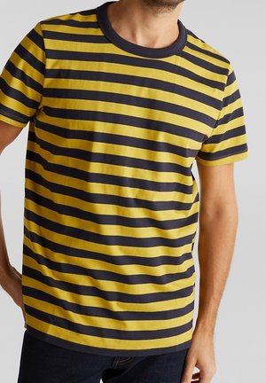 Homme portant un T-shirt à manches courtes à rayures horizontales jaunes et noires avec un col noir, associé à un pantalon foncé.
