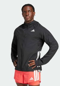 Chaqueta atlética negra con cremallera en el frente, ligeramente brillante, con detalles de tres franjas blancas a lo largo de los lados. Incluye capucha y cuello alzado.