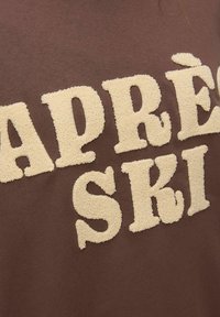 Sudadera marrón con letras texturizadas en beige claro que forman la frase "APRÈS SKI". La tela parece suave con un corte informal.