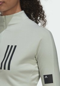 Primer plano de una persona con una chaqueta de cremallera gris claro que tiene tres rayas negras en el pecho y un parche del logo de Adidas en la manga.