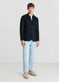 Blazer marine avec boutons dorés, deux poches avant, associé à un jean bleu clair et des chaussures beiges à lacets. Tissu lisse, forme structurée.