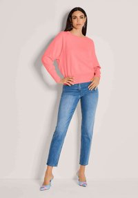Roze gebreide trui met brede mouwen, lichtblauwe jeans en lichtblauwe hakken met een roze en witte accentdetail. Neutrale achtergrond.
