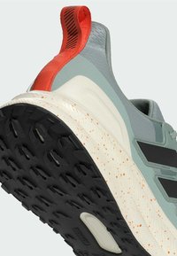 Teniska s svetlozeleným zvrškom, čiernymi pruhmi, červenou pätkou a štruktúrovanou čiernou podrážkou s oranžovými bodkami.