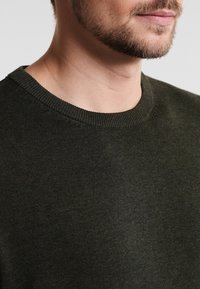 Tmavě zelený svetr s kulatým výstřihem a žebrovaným límcem. Materiál se zdá být měkký, s hladkou texturou a minimálními detailem šití.