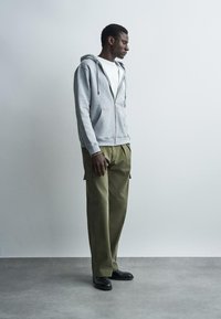 Młody mężczyzna w jasnoszarej bluzie na zamek, białej koszulce, oliwkowozielonych spodniach typu cargo i czarnych butach stoi na tle jednolitej szarej ściany.