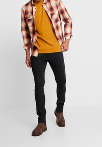 Camisa de franela a cuadros en rojo, naranja y blanco, superpuesta sobre una camiseta mostaza. Jeans negros ajustados y botas de cuero marrón.
