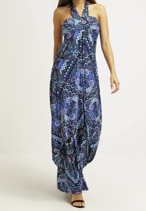 Woman wearing a sleeveless, blue and purple patterned halter-neck maxi dress with black open-toe heels, standing against a plain background.