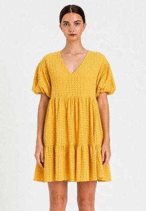 Donna che indossa un vestito corto giallo strutturato con maniche a sbuffo e scollo a V profondo, in piedi davanti a uno sfondo chiaro uniforme.