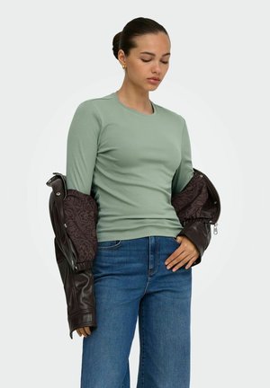 Femme aux cheveux foncés en chignon, portant une chemise vert clair à manches longues et un jean bleu, tenant une veste en cuir marron foncé à moitié sur les bras.