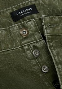 Maslinasto zelena traper tkanina s blagim sjajem, s crnom etiketom na kojoj piše "JACK & JONES" i dva metalna dugmeta na kopčanje.