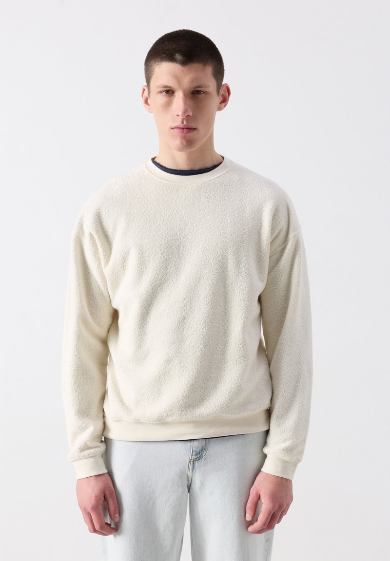 Mladý muž s krátkými vlasy, oblečený v krémovém texturovaném svetru s dlouhým rukávem a světle modrých džínách, stojící před jednobarevným bílým pozadím.