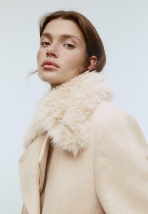 Jasno beżowy płaszcz z puszystym, teksturowanym kołnierzem z futra syntetycznego. Projekt obejmuje dopasowaną sylwetkę i ukryte kieszenie boczne.