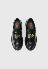 Black leather loafers with a glossy finish, featuring a gold "LOVE MOSCHINO" strap, cushioned soles, and heart-patterned insoles.