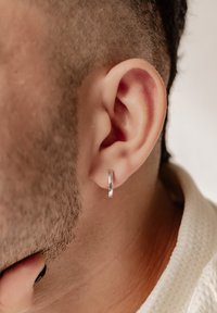 Pendiente de aro plateado exhibido en la oreja, con una superficie pulida y un diseño circular. Se puede ver el cabello corto y la textura de la piel cercana.