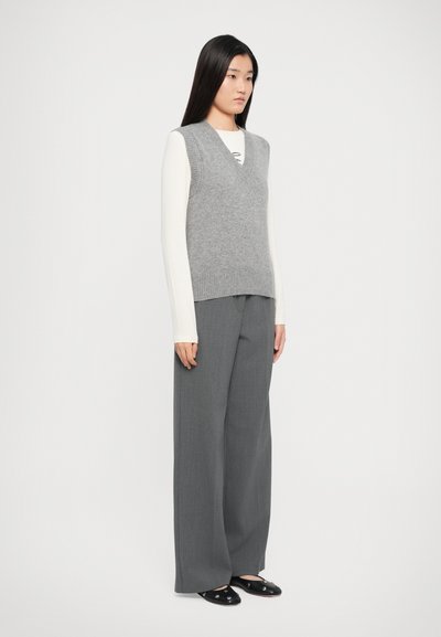 Woman standing wearing a grey sleeveless sweater vest over a white long-sleeve top, gray wide-leg pants, and black studded flats.