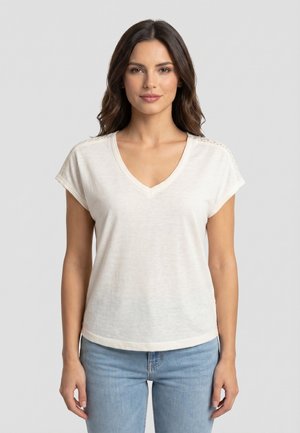 Žena s hnědými vlasy, oblečená v bílém tričku s véčkovým výstřihem a krátkými rukávy a světlemodrých džínách, stojící na jednobarevném světle šedém pozadí.